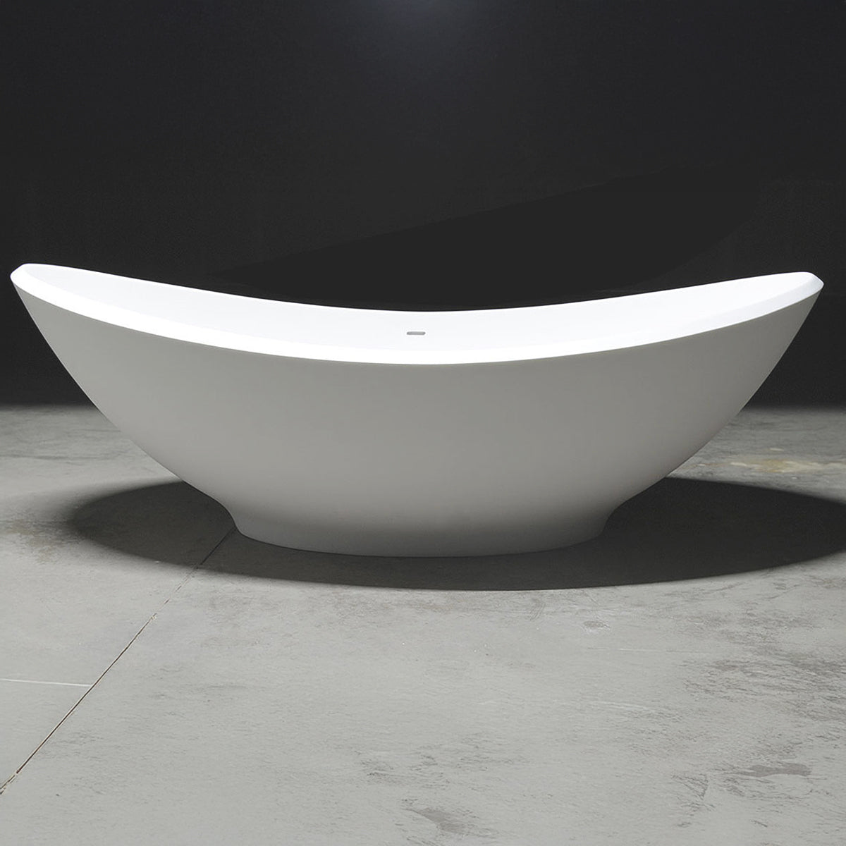 Magnus Hartland solid surface stone resin freestanding bathtub in matte white — studio shot of the double-slipper tub, with its dipping center rim and raised sweeping ends, on a marble floor against a dark background.